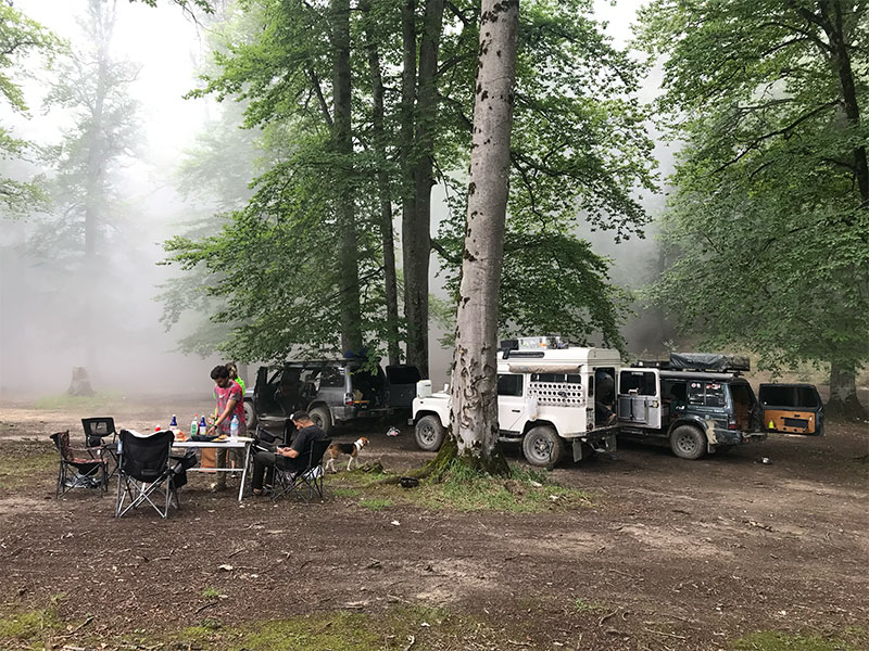 Ein Overlanding-Camp mit drei Fahrzeugen, bei dem bereits ein Tisch mitsamt Stühlen aufgebaut wurden. Die Fahrzeuge stehen im Hintergrund im nebligen und gruenen Wald.