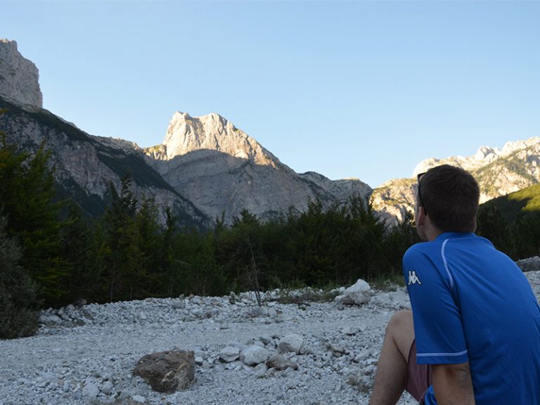 Kevin blickt in steiniger Landschaft hinauf zum sonnenbeleuchteten Gipfel des Berg Arapi im albanischen Theth Nationalpark.