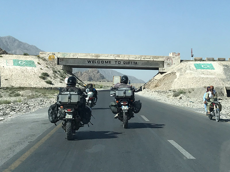 Zwei Motorradreisende folgen dem Polizeimotorrad unter einer Brücke mit der Aufschrift Quetta. Dies ist der Beginn der belutschischen Provinzhauptstadt Quetta in Pakistan.